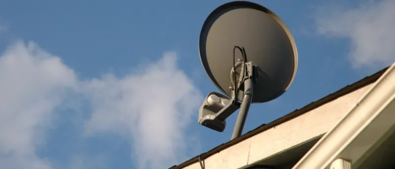 A gray satellite dish mounted on the edge of a light-colored roof, set against a bright blue sky with white clouds.